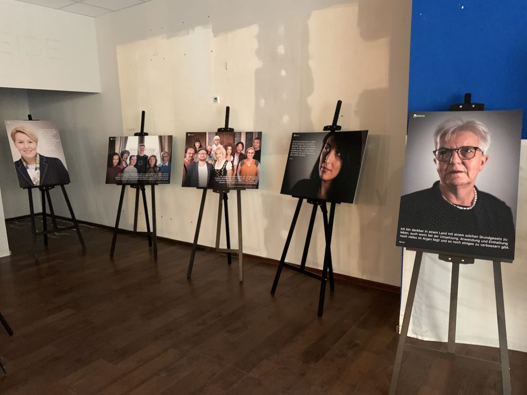 Kunst-Ausstellung Gesichter der Demokratie: Menschen sind einzeln oder in Gruppen auf Leinwänden abgebildet. Ein Zitat der Menschen zum Thema Demokratie steht daneben.